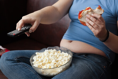 Meisje Dat Op De Bank Tv Zit Te Kijken Met Een Bak Popcorn Op Schoot En Een Broodje In Haar Hand Als Voorbeeld Van Slechte Afvalgewoontes