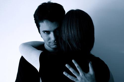 Een man kijkt spottend naar de camera terwijl hij een vrouw omhelst