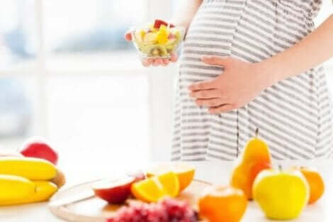 Een zwangere vrouw staat achter een tafel met vruchten en heeft een bakje fruit in haar hand